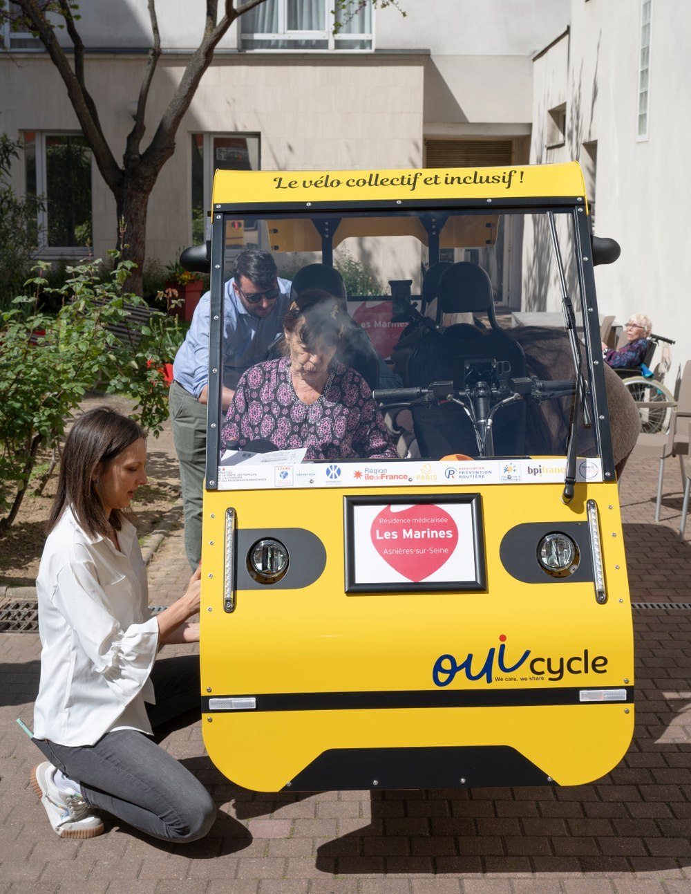 Le OuiCycle présenté dans la cour d’une résidence médicalisée : une résidente installée à bord, l’accompagnante et un proche à leurs côtés.