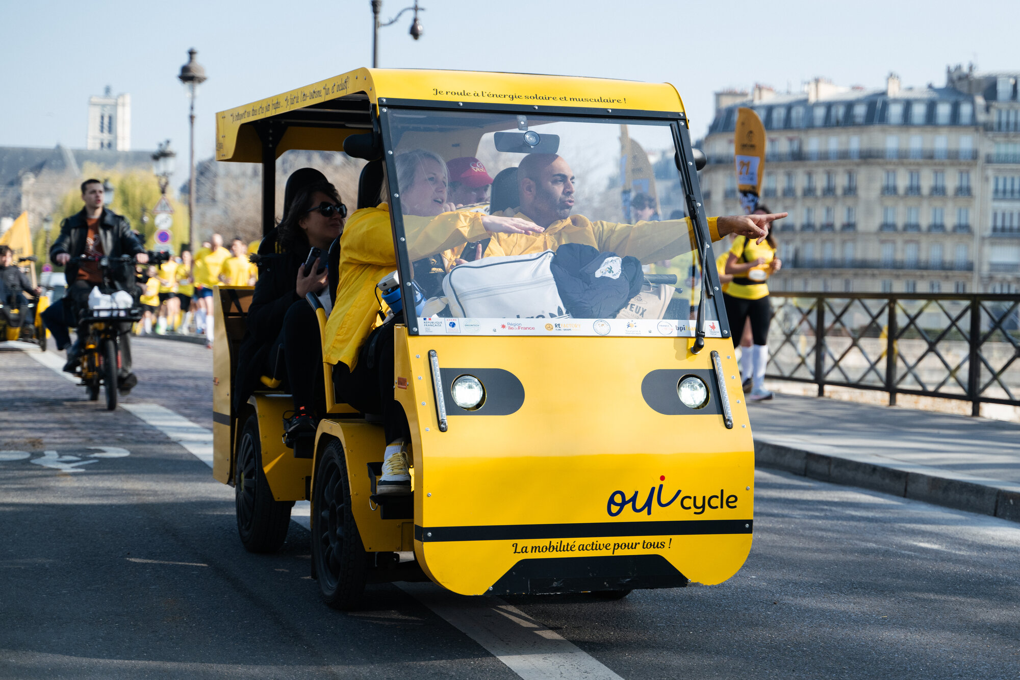 Le OuiCycle jaune en sortie sur un pont parisien : le conducteur-animateur montre l'horizon, des passagers souriants à bord.