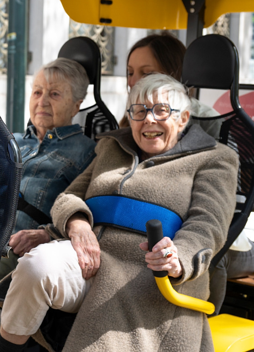 Deux résidentes installées dans le vélo collectif jaune, l'une éclate de rire, accompagnées de leur animatrice : la joie comme indicateur thérapeutique.