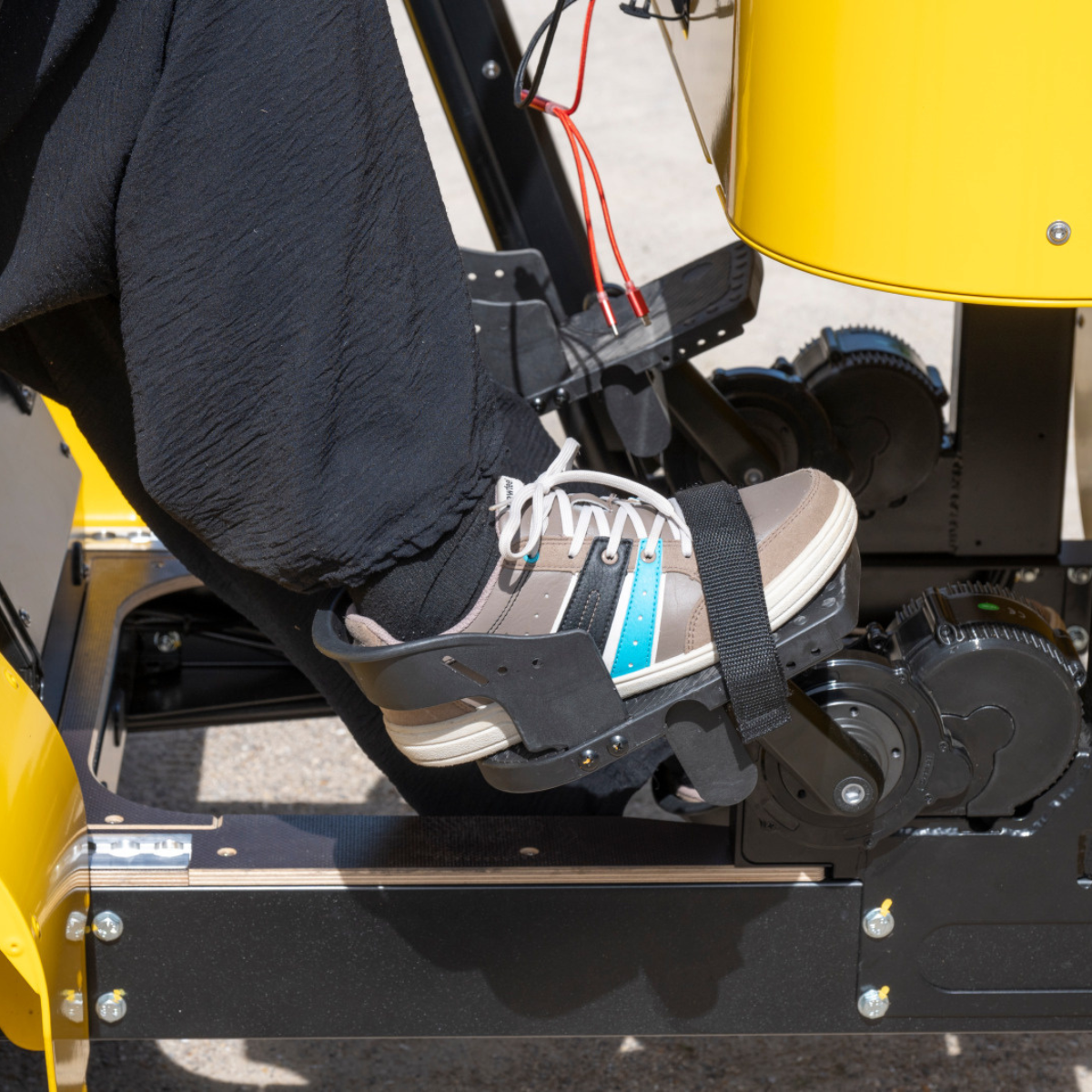 Gros plan sur le pied d'un passager, sanglé et maintenu sur la pédale du vélo collectif jaune Les Roues Solidaires pour un pédalage sécurisé.